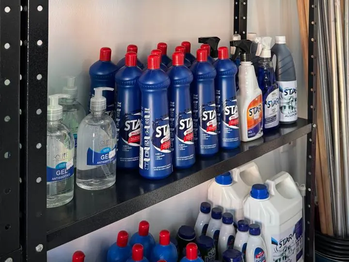 Prateleira com frascos azuis de limpador multiuso, álcool em gel e galões de produtos de limpeza