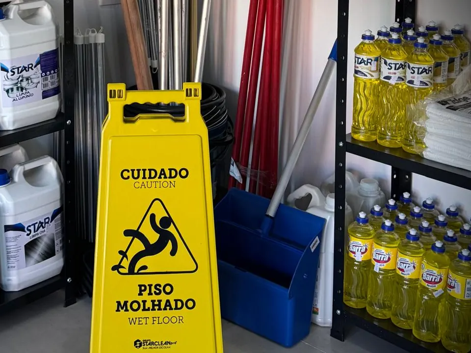 Placa amarela de piso molhado ao lado de balde com mop, cabos de vassoura e galões de limpador
